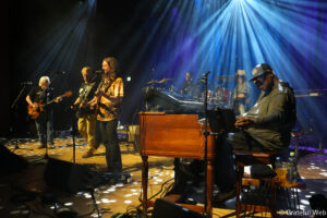 Live band on stage performing under blue spotlight, with guitarists, a keyboard player, and a drummer visible in the background.
