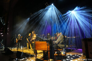 Band performing on a dimly lit stage with blue spotlight beams and multiple musicians visible (guitarists, keyboard, drums).
