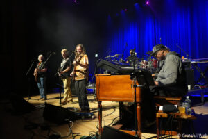 Live band on stage performing with guitarists, a keyboard player, and a drummer under blue stage lights; cables and gear on the floor in front.