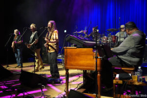 A group of musicians performing on a stage with guitars and a drum set under purple and blue stage lighting during a live concert.