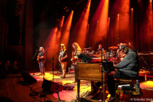 Live concert scene with a band performing on a dimly lit stage bathed in orange and red spotlights; guitarists, keyboardist, and drummer visible.