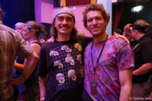 Two friends pose together at a lively party, one in a skull-print black shirt and the other in a purple tie-dye shirt, smiling with arms around each other.