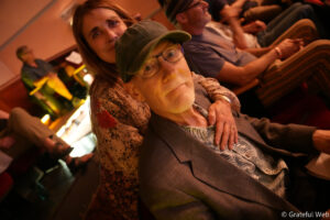 A smiling elderly man in a green cap and glasses being hugged from behind by a woman in a floral blouse at a warmly lit event venue.