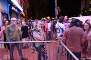 Crowd of people at a street festival at night, smiling and chatting behind metal barriers with neon lights in the background.