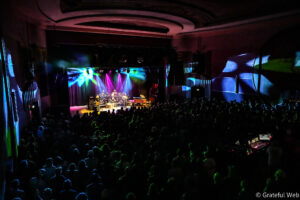 A live concert on a brightly lit stage with multiple musicians performing as colorful lights wash over the performers and the audience fills the dark theater.