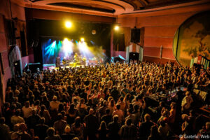 Large crowd watches a live band on stage in a theater with colorful spotlights overhead.