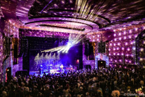 Large indoor concert with a band performing on stage, purple and white lighting, and a packed audience watching.