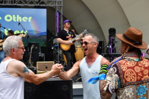 Two men in a crowd fist-bump in front of a live band on stage outdoors at a festival stage backdrop behind them showing words and a rainbow image