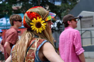 Woman in a colorful, bundled hat with a bright sunflower, denim vest with bead embroidery, at an outdoor festival.