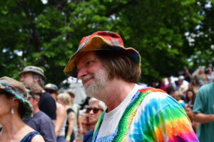 Man with gray beard wearing a colorful tie-dye shirt and a rainbow bucket hat at an outdoor festival.