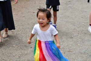 Young girl wearing a white shirt and a rainbow tutu walking on a gravel path, with adults' legs nearby.