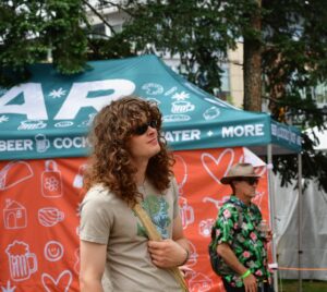 Man with long curly hair and sunglasses, wearing a beige T-shirt and crossbody bag, at an outdoor beer festival in front of a teal tent shift-signaled canopy.