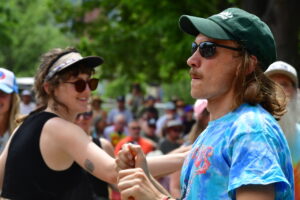 Two people dancing hand in hand at an outdoor event with a crowd in the background; man wearing a green cap and sunglasses, woman wearing a visor and round sunglasses smiling at him