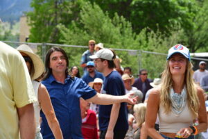 Crowd at an outdoor event, with a smiling woman wearing a blue cap on the right amid trees and a sunny day.
