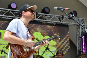 Guitarist on stage wearing a cap and sunglasses, playing a brown electric guitar with a microphone nearby and colorful stage backdrop.