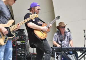 Guitarist in a purple cap and black shirt plays an orange electric guitar on a stage, with a keyboardist wearing sunglasses and a hat in the background.