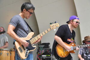Two male musicians perform on an outdoor stage: left guitarist bass-playing in a gray shirt and sunglasses, right guitarist in a black shirt with a sunburst guitar and purple cap sunglasses.