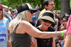 Two women at an outdoor festival smiling and chatting, one wearing a black visor and sunglasses on a sunny day.