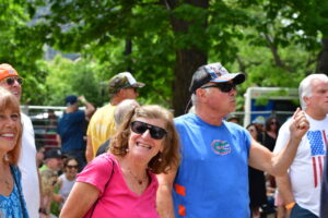 Smiling group at an outdoor festival; a woman in pink foreground with sunglasses beside a man in a blue Florida Gators shirt and cap.