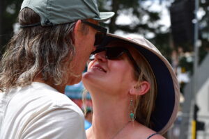 Couple sharing a kiss outdoors, both wearing hats and sunglasses.",