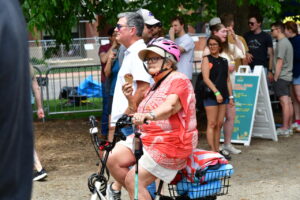 Older woman wearing a pink helmet rides a small bicycle with a basket, eating an ice cream cone at an outdoor event.