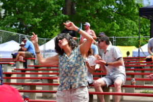 Woman in a floral sleeveless blouse dances with arms raised at an outdoor festival, with spectators on red bleachers behind her.