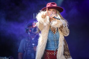 Female singer on stage wearing a burgundy hat and furry coat, pointing toward the audience while holding a microphone against a purple-lit background.