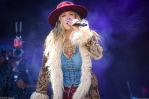 Female singer wearing a maroon hat and fur coat, singing into a microphone on a smoky stage with purple lighting.