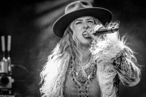 Female singer performing on stage, wearing a wide-brim hat and a fluffy jacket, microphone in hand and singing intensity evident.