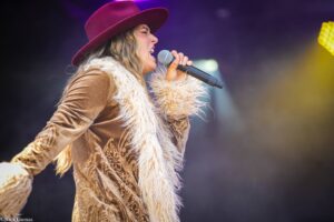 Female singer wearing a maroon wide-brim hat and furry beige vest performs into a handheld microphone on stage.