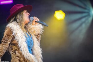 Female singer in a red wide-brim hat belts into a microphone on stage, wearing a furry coat and denim outfit with colorful stage lights in the background