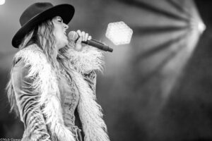 Female singer wearing a wide-brim hat and fluffy jacket singing into a handheld microphone on stage, black-and-white image.