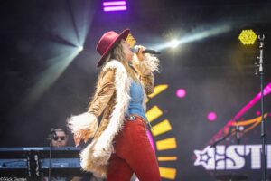 Female singer on stage singing into a microphone, wearing a red hat and furry vest over denim, with bright stage lights in the background.