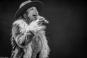 Female singer wearing a wide-brim hat and fringed jacket performs into a microphone on stage (black and white).