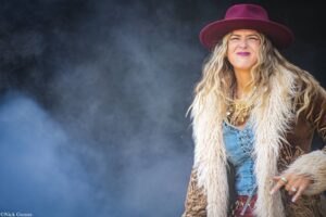 Smiling woman in a burgundy wide‑brim hat, denim jacket, and furry coat stands in a smoky blue background, looking toward the camera edge.