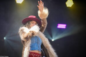 Female performer on stage waving to the crowd, wearing a red hat, denim vest, and furry coat under colorful stage lights.