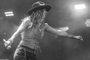 Female singer on stage wearing a wide-brim hat, denim vest, and jewelry, singing into a handheld microphone with arms outstretched.