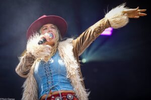 Female singer performing on stage, holding a microphone with one hand and raising the other, wearing a burgundy hat, denim top, and faux-fur jacket.