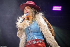 Female singer wearing a red hat and denim outfit, performing into a microphone on stage, with a furry coat flowing around her.