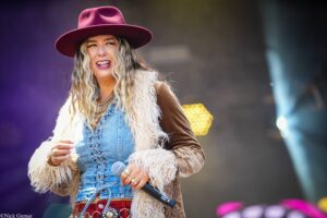 Female singer on stage wearing a burgundy wide-brim hat, denim vest, and furry coat, holding a microphone.