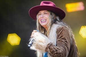 Woman laughing on stage in a burgundy hat and brown jacket, holding a microphone with yellow stage lights in the background.