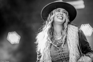 Joyful woman wearing a wide-brim hat and denim jacket on stage, smiling widely with layered jewelry and a furry vest.