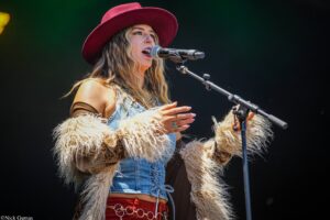Female singer performing on stage, wearing a wide red hat and denim vest, singing into a microphone.