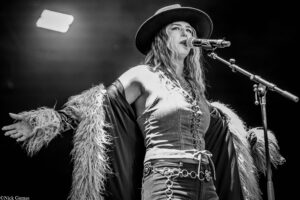 Black-and-white photo of a female singer performing on stage, wearing a wide-brimmed hat, denim outfit, and a fur-trimmed cape, singing into a microphone with arms spread wide.
