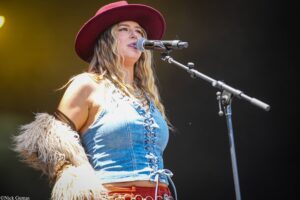 Female singer wearing a red wide-brim hat and denim outfit performs on stage into a microphone.