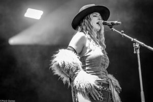 Female performer wearing a wide-brim hat and denim, singing into a microphone on stage.