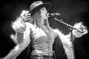 Female singer wearing a wide-brim hat and fringed denim top performs into a microphone on stage.