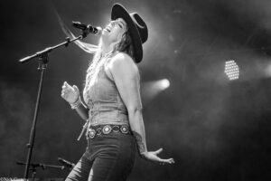 Female singer wearing a wide-brim hat and denim outfit, singing into a microphone on stage.