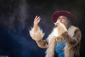 Female singer wearing a burgundy wide-brim hat and fluffy coat, holding a microphone on stage.