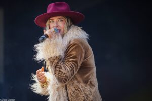 Female singer performing on stage, wearing a burgundy wide-brim hat and a fringed fur coat while holding a microphone.